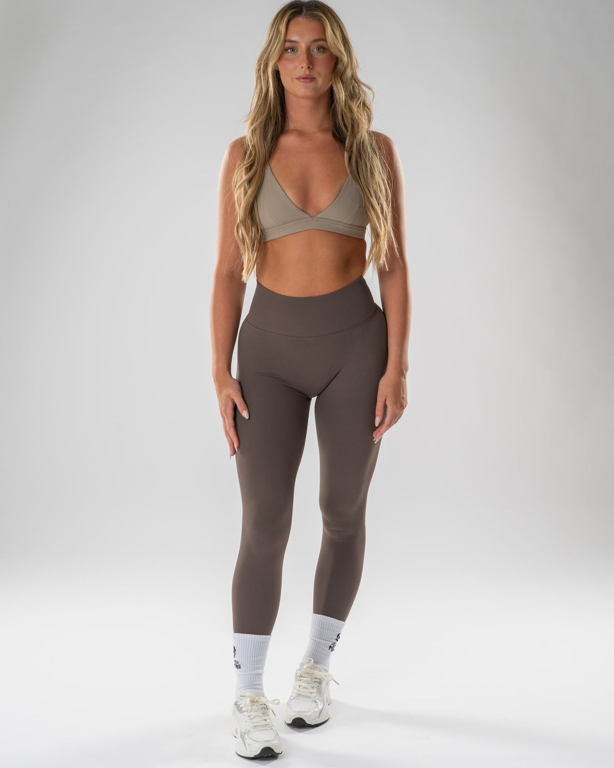 A woman modeling high-waisted taupe-brown leggings and a matching beige sports bra against a light gray studio background.