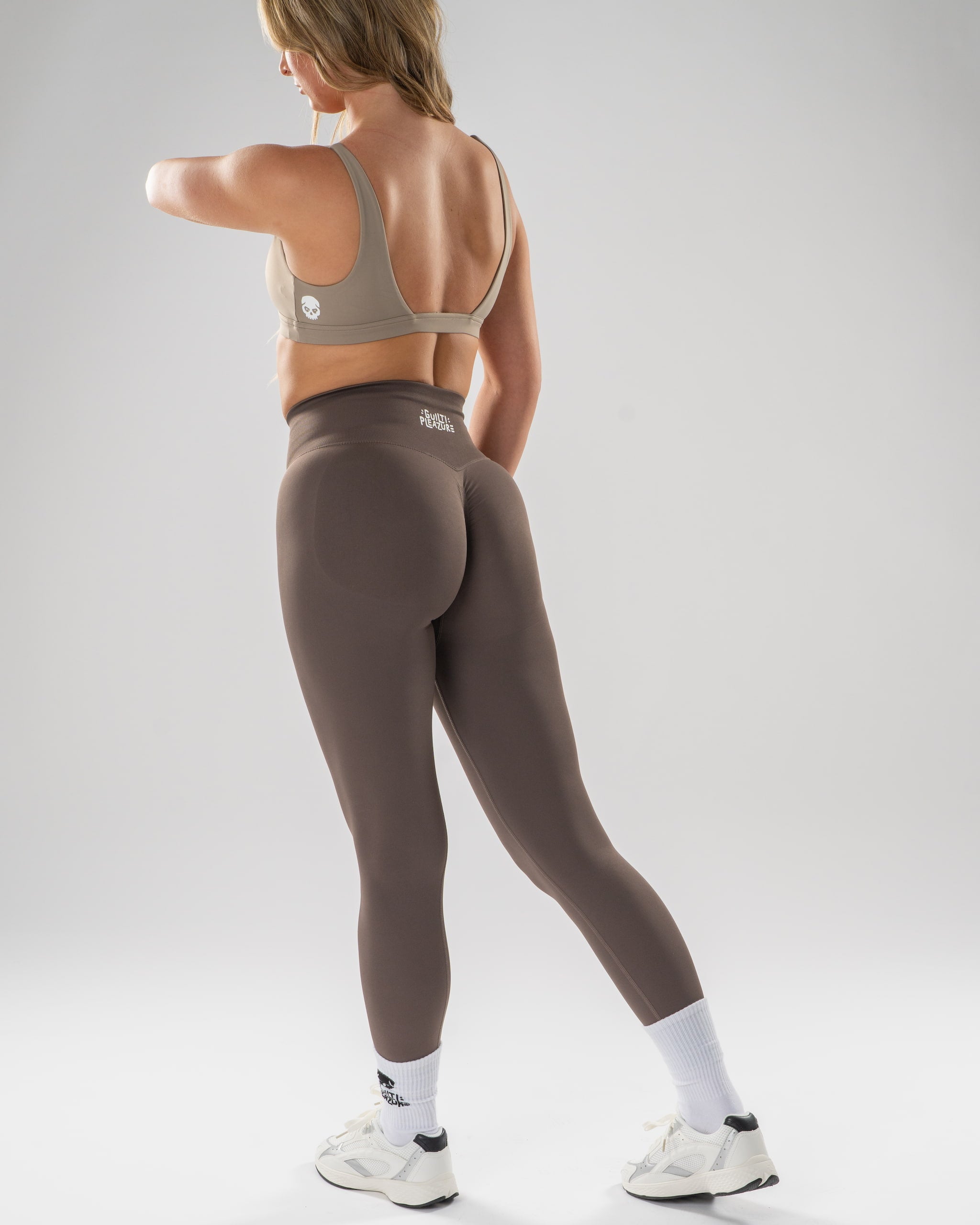 A woman wearing a taupe sports bra and matching high-waisted leggings, shown from the back in a studio setting with a light gray background.