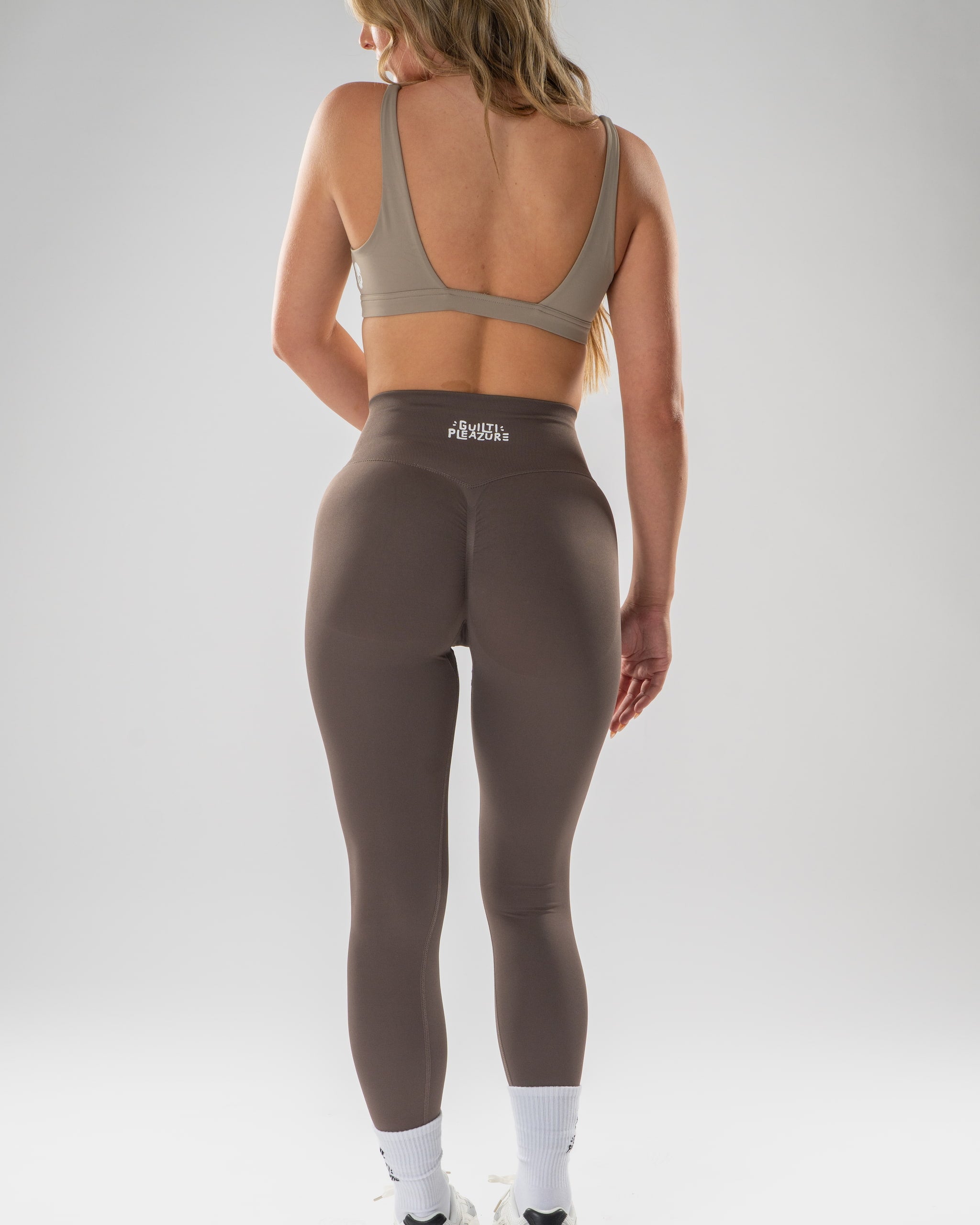 A woman is shown from behind wearing high-waisted athletic leggings and a matching sports bra, standing against a light gray studio background.