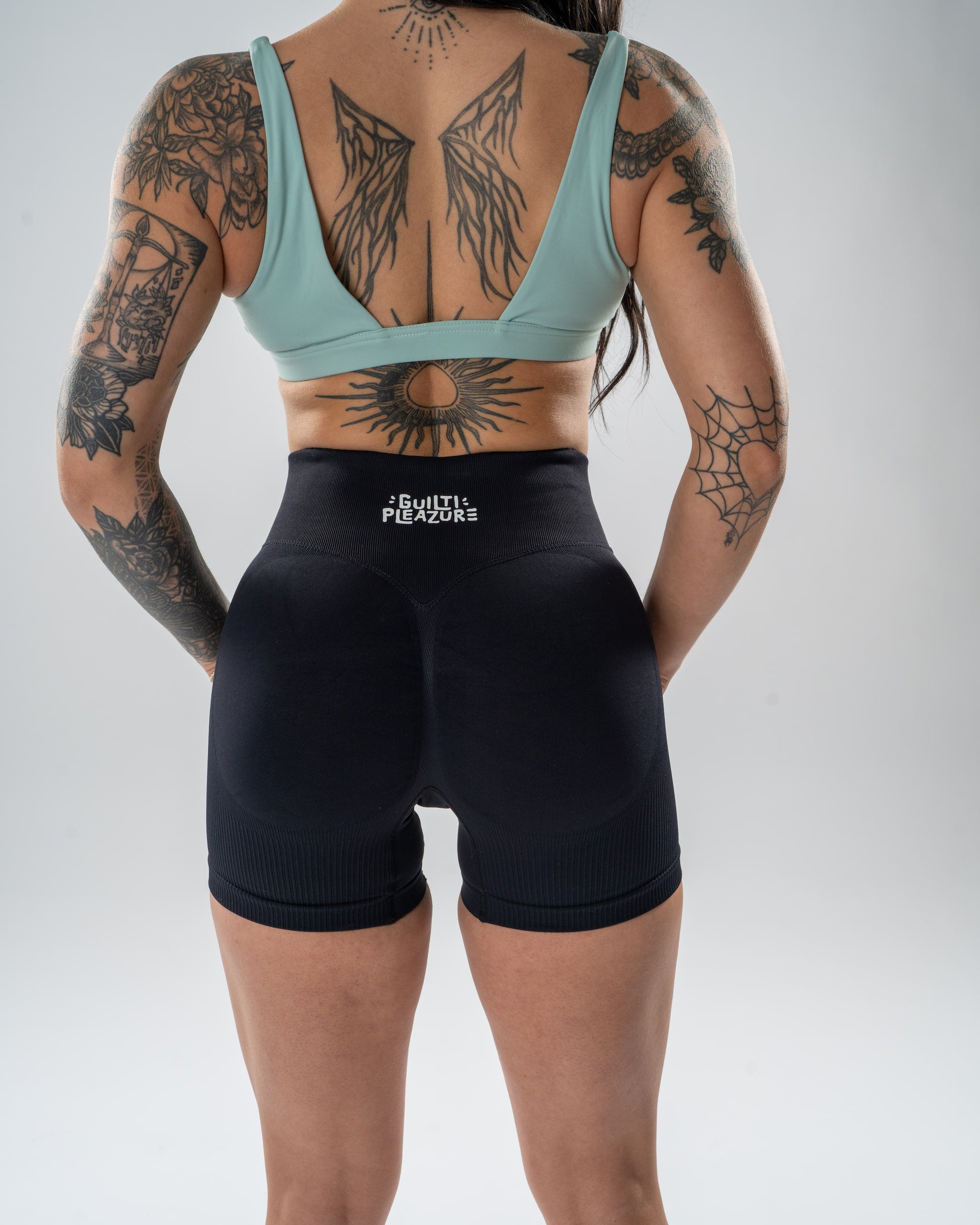 Rear view of a tattooed woman wearing black athletic shorts with a white 'BREATHE FITNESS' logo on the back waistband, paired with a light blue sports bra, against a light gray studio background.
