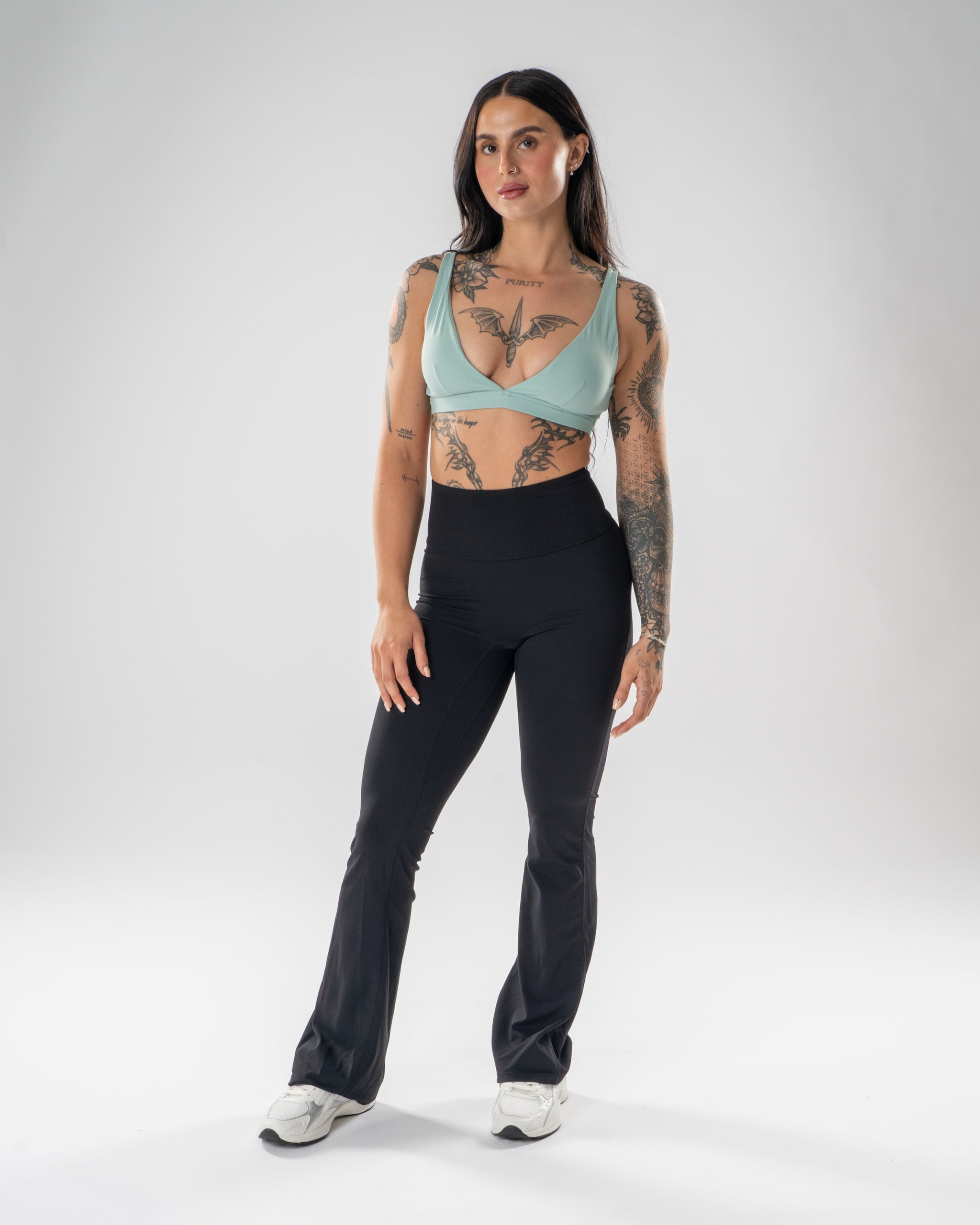 A woman modeling a two-piece activewear set consisting of a light sage green scoop-neck sports bra and black high-waisted flared leggings, standing against a light gray studio background.