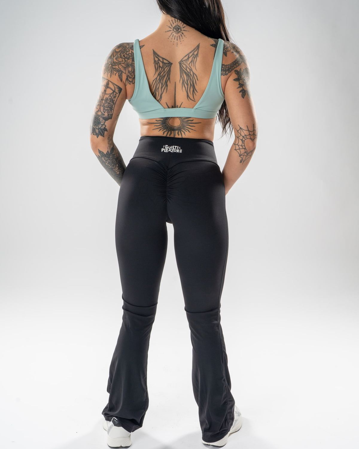 A woman viewed from the back wearing black high-waisted flared leggings and a mint green sports bra, standing against a light gray studio background.