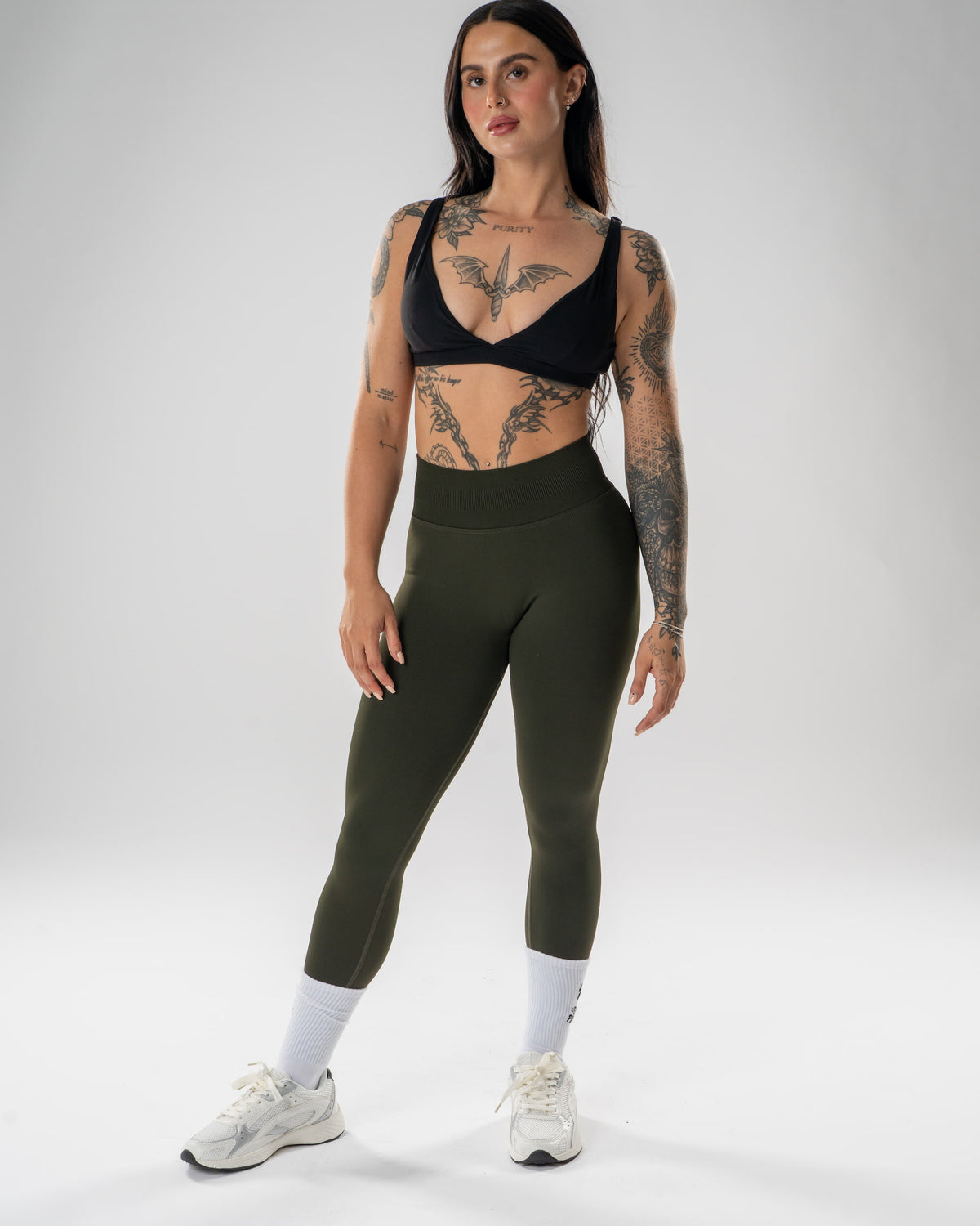 A woman modeling high-waisted olive green athletic leggings, a black sports bra, white socks, and white sneakers against a light gray studio background.