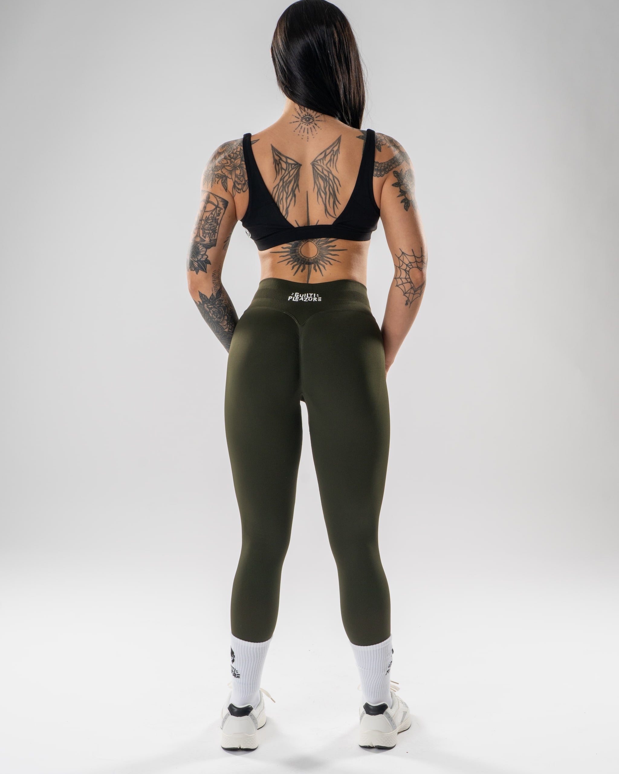 A woman stands with her back to the camera wearing olive green high-waisted leggings, a black sports bra, white socks, and white sneakers on a light gray studio background. The brand name 'altea' is visible on the waistband of the leggings.