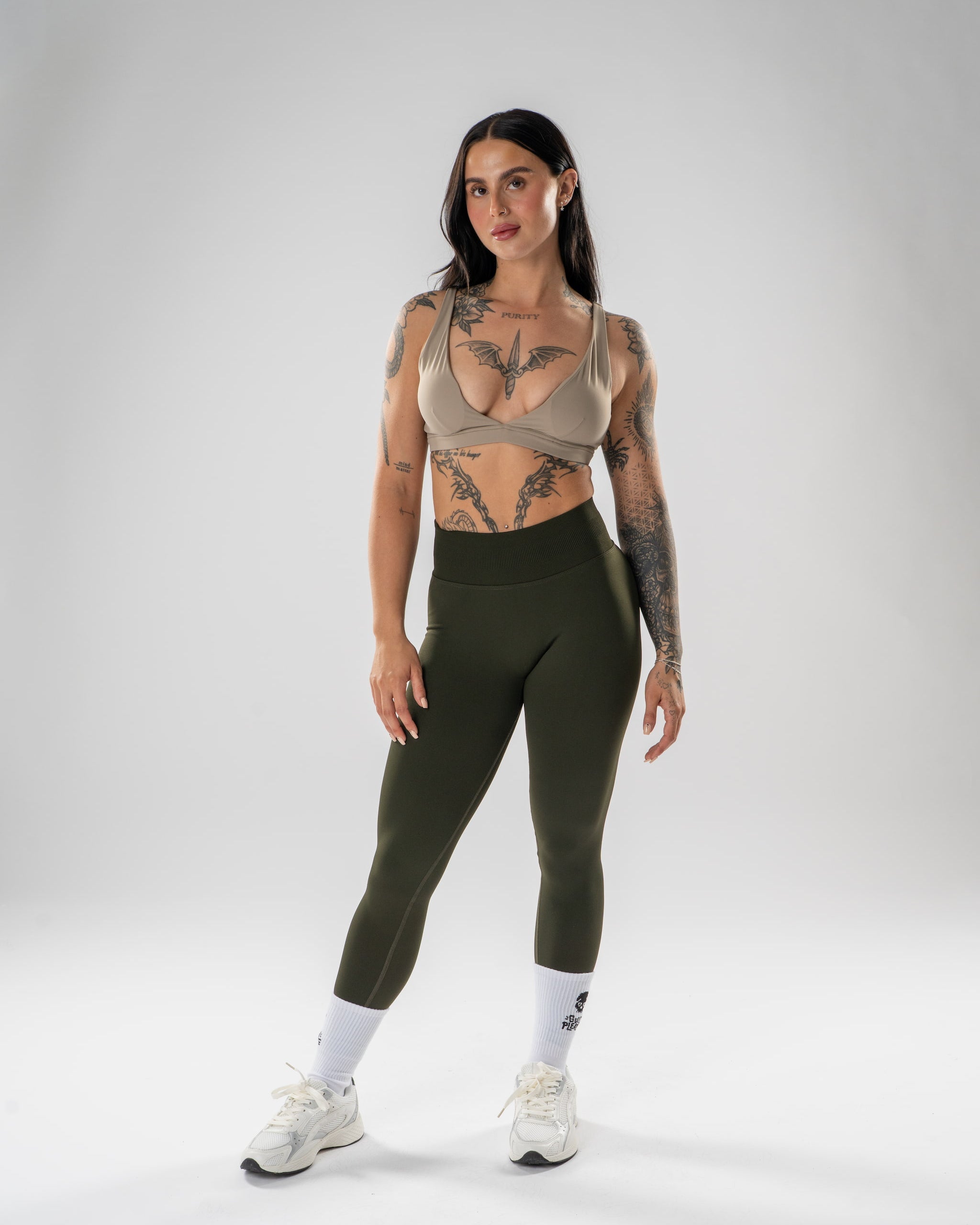 A woman modeling a matching athletic set consisting of an olive green high-waisted leggings and a taupe-beige sports bra, paired with white socks and white sneakers, against a light gray studio background.
