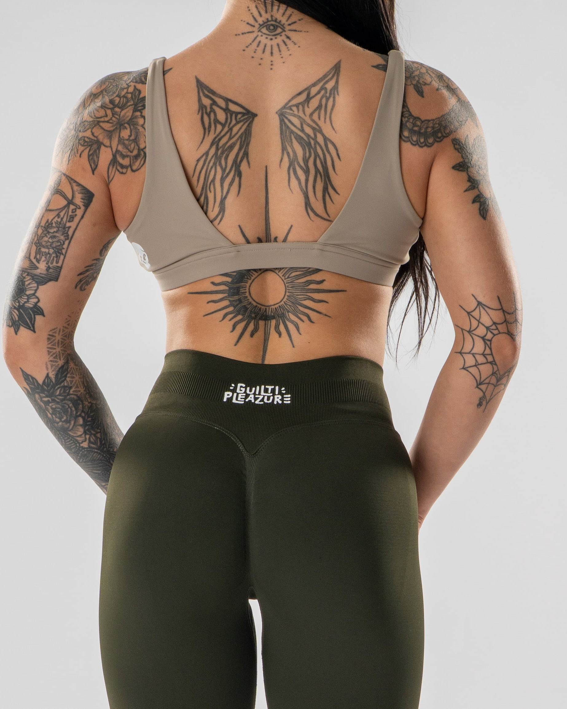 Rear view of a tattooed woman wearing a taupe sports bra and olive green leggings with the brand name 'EXCELLENT FUSION' on the waistband, against a light gray studio background.