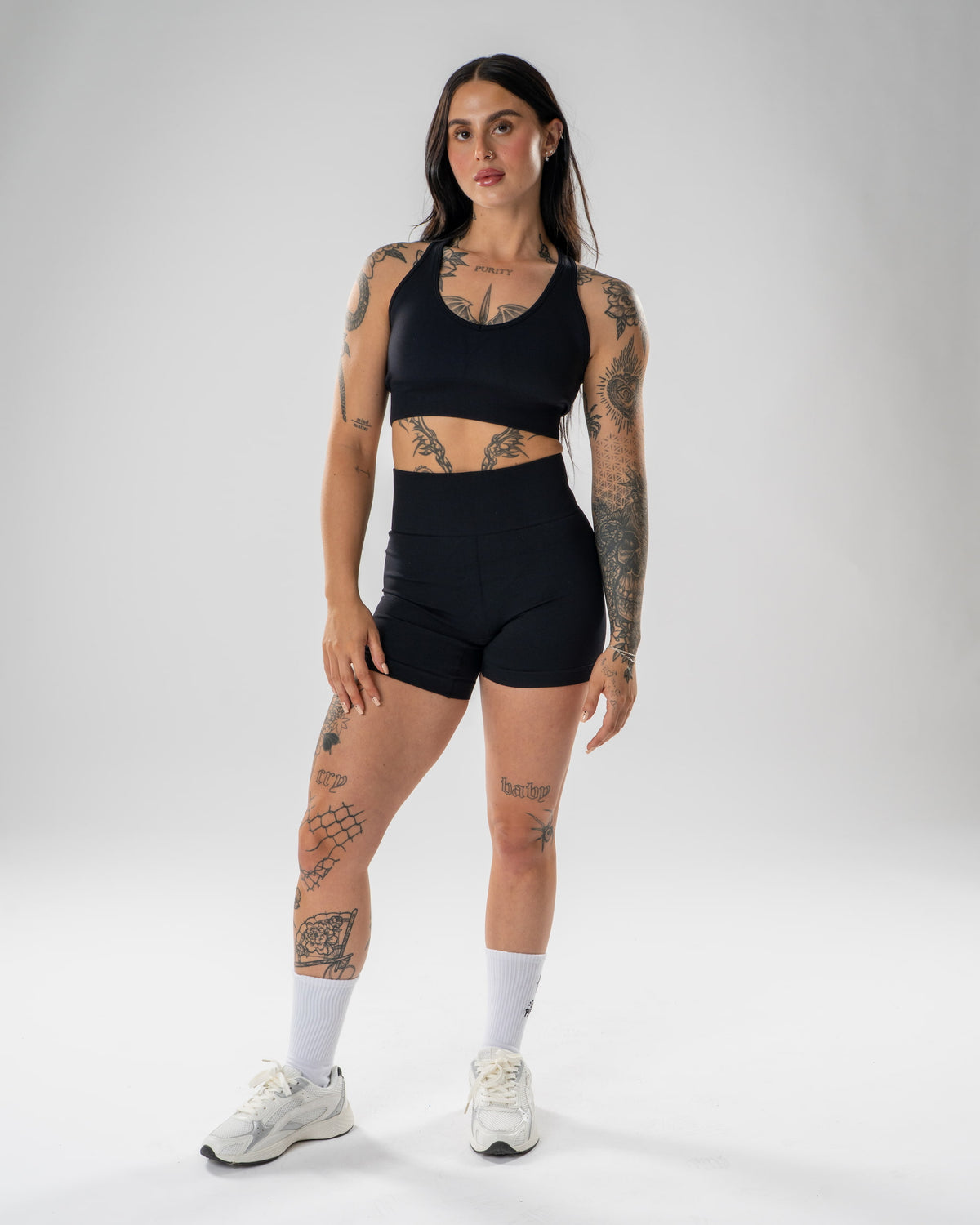 A woman modeling a matching black activewear set consisting of a sports bra and high-waisted shorts, paired with white socks and white sneakers, against a light gray studio background.