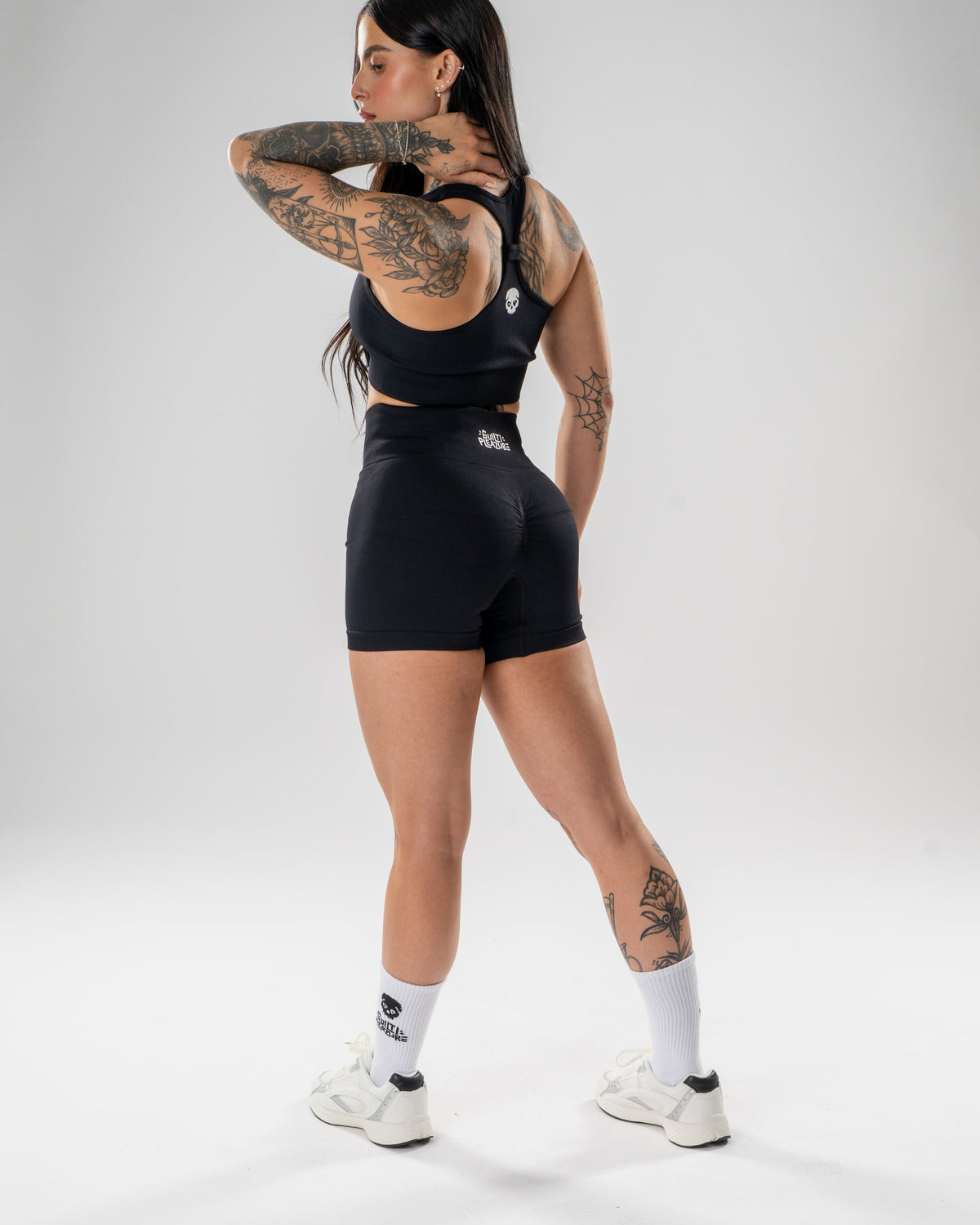 A woman modeling a black athletic set consisting of a sports bra and matching shorts, shown from a rear three-quarter angle against a light gray studio background.