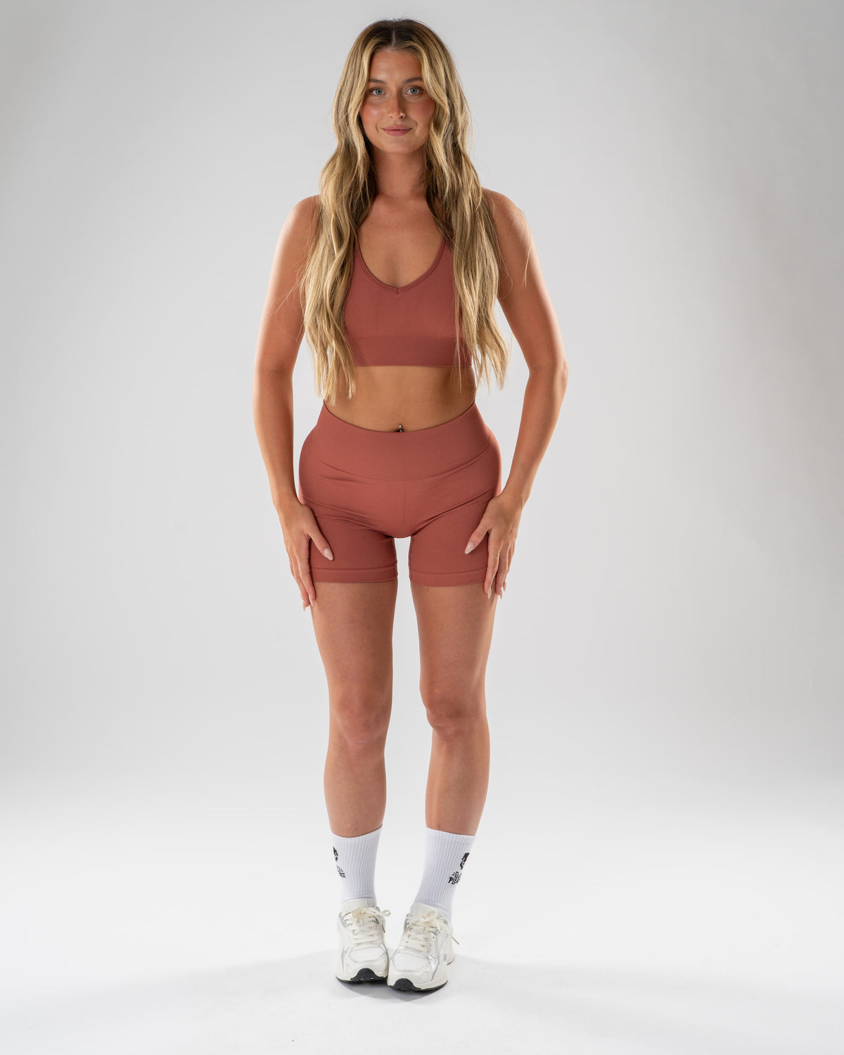 A woman modeling a matching terracotta-colored activewear set consisting of a scoop-neck sports bra and high-waisted biker shorts, paired with white crew socks and white sneakers, against a light gray studio background.