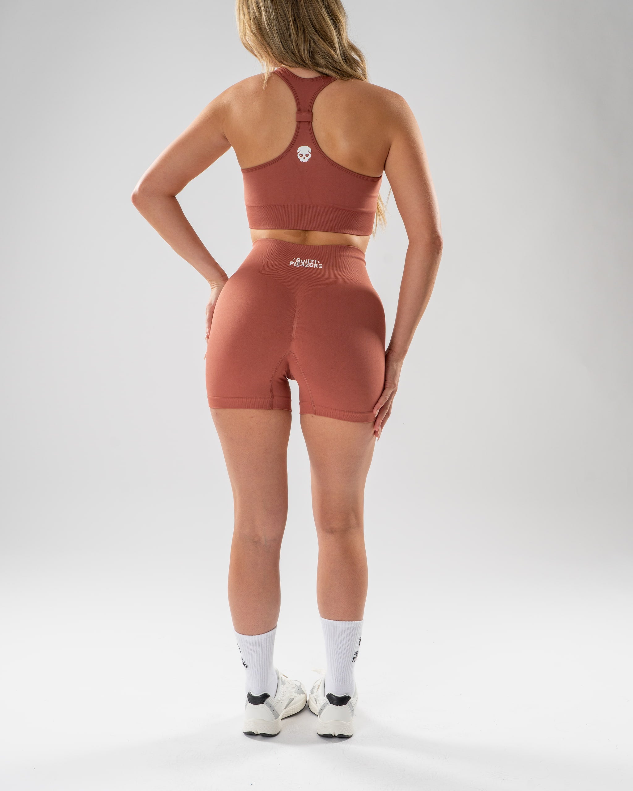 A woman stands with her back to the camera wearing a matching terracotta-colored athletic set consisting of a sports bra and high-waisted shorts, paired with white socks and sneakers, against a light gray studio background.