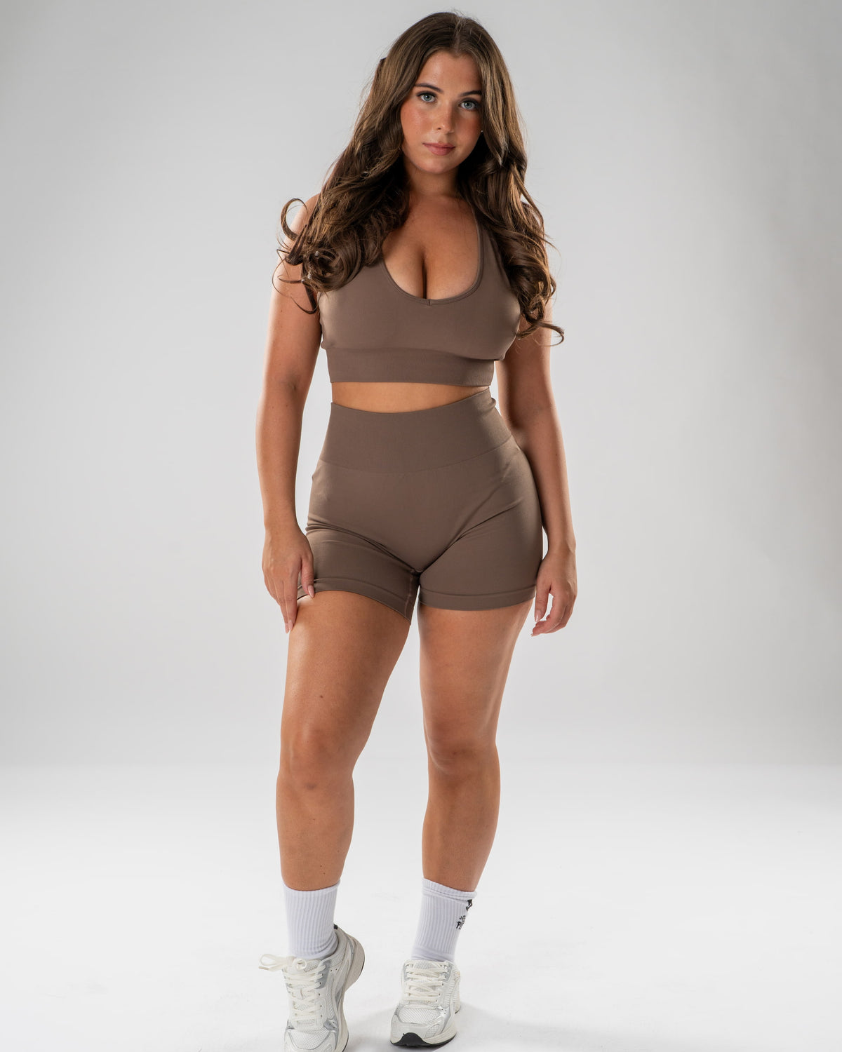 A woman modeling a matching taupe brown activewear set consisting of a scoop-neck sports bra and high-waisted shorts, paired with white socks and white sneakers, against a light gray studio background.