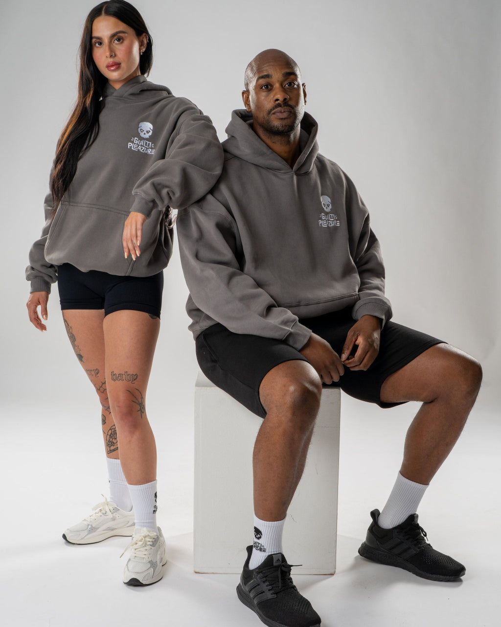 A man and a woman modeling matching dark gray hoodies with a white shield logo on the chest, paired with black athletic shorts and white or black sneakers, posed against a light gray studio background.