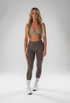 A woman modeling high-waisted taupe-brown leggings and a matching beige sports bra against a light gray studio background.