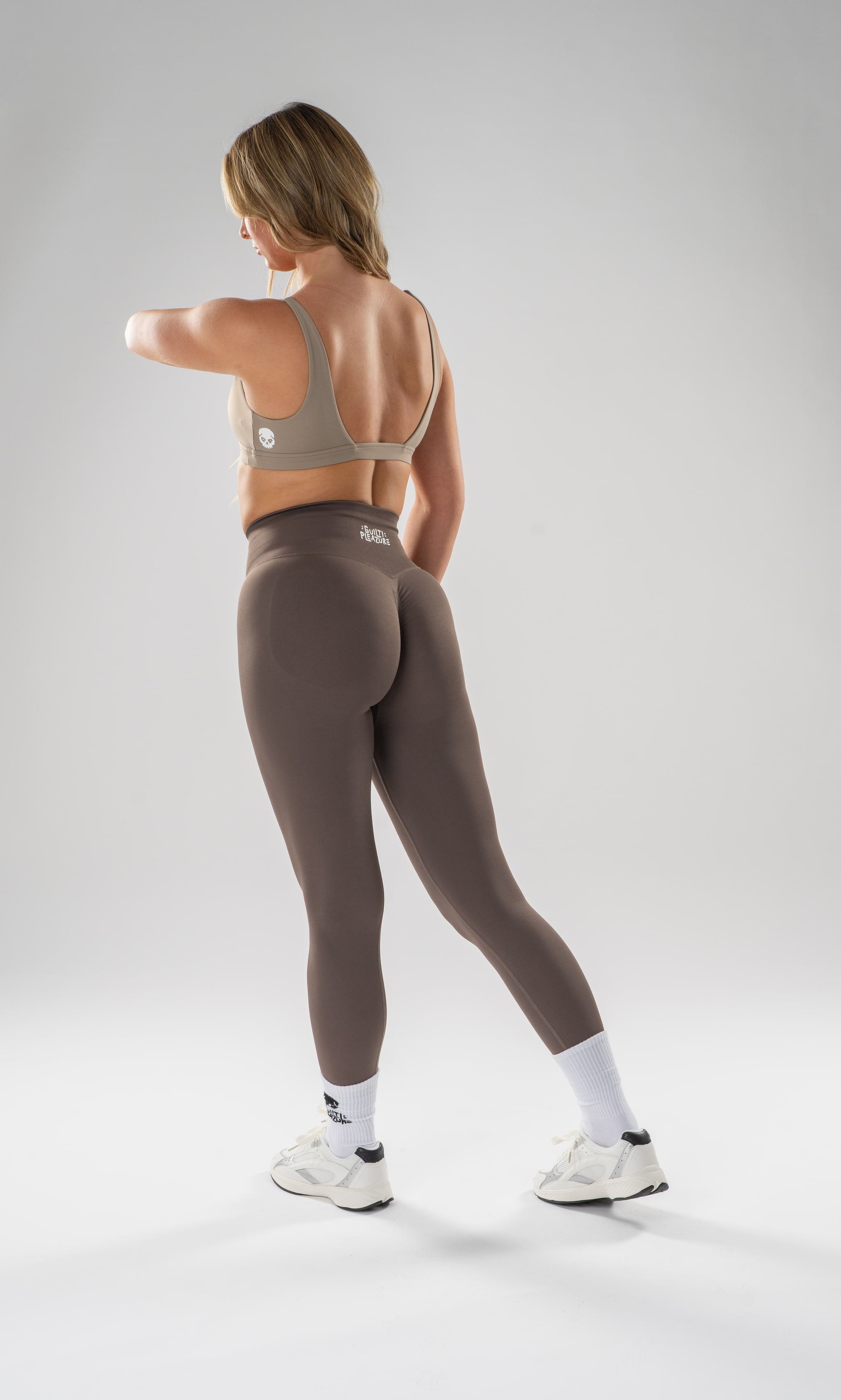 A woman wearing a taupe sports bra and matching high-waisted leggings, shown from the back in a studio setting with a light gray background.