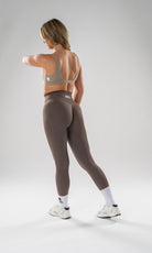 A woman wearing a taupe sports bra and matching high-waisted leggings, shown from the back in a studio setting with a light gray background.