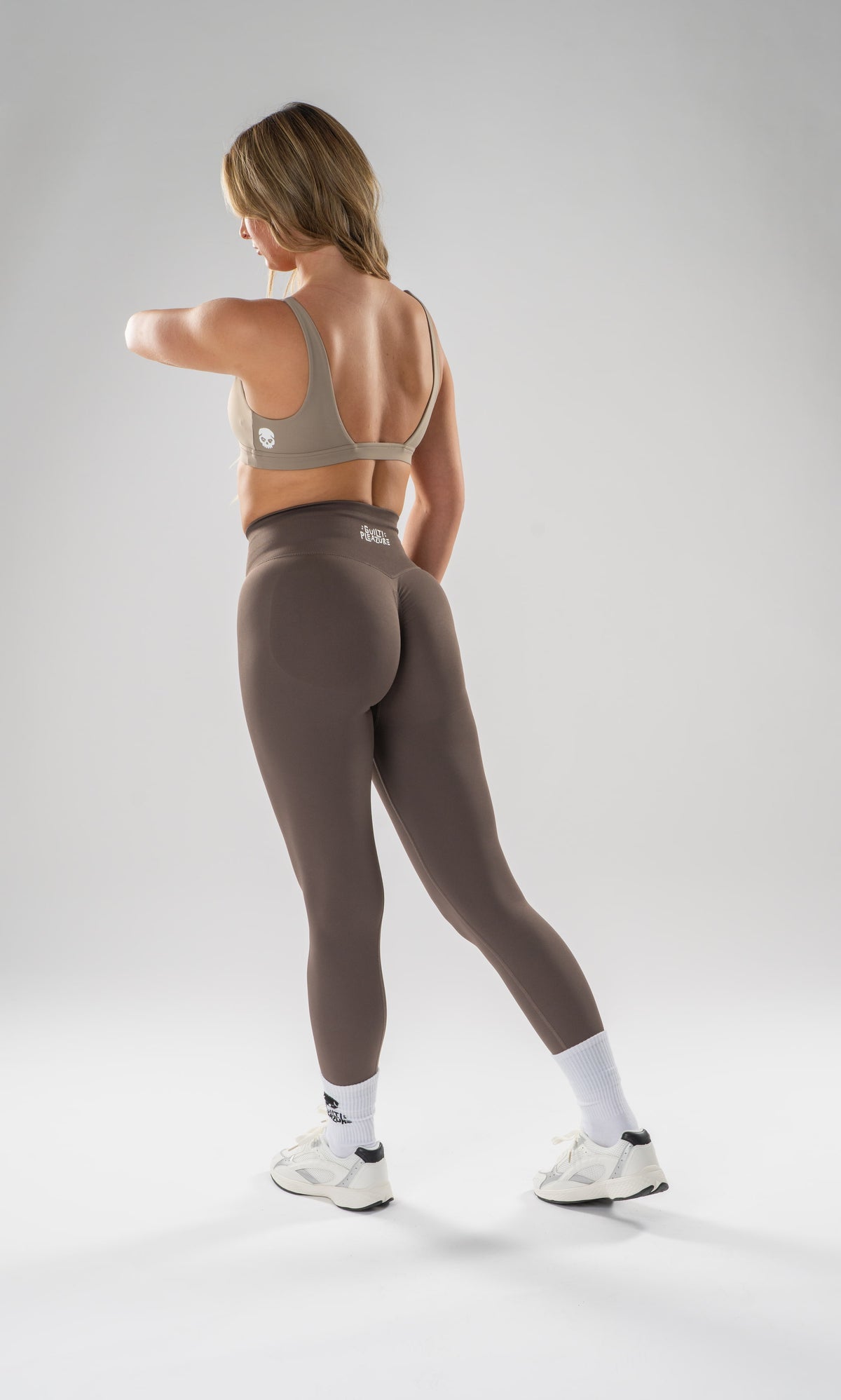 A woman wearing a taupe sports bra and matching high-waisted leggings, shown from the back in a studio setting with a light gray background.
