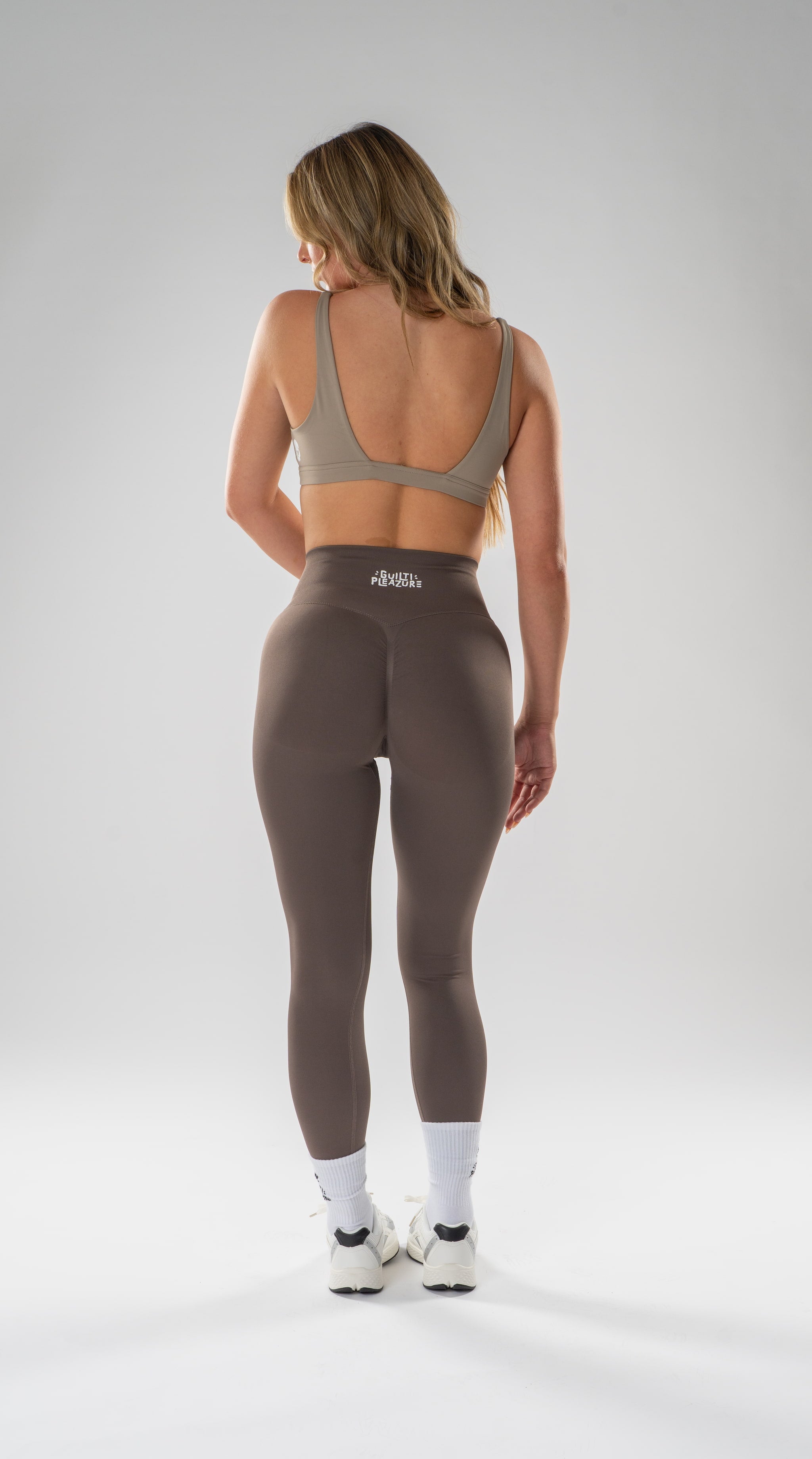 A woman is shown from behind wearing high-waisted athletic leggings and a matching sports bra, standing against a light gray studio background.