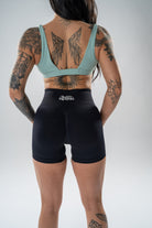 Rear view of a tattooed woman wearing black athletic shorts with a white 'BREATHE FITNESS' logo on the back waistband, paired with a light blue sports bra, against a light gray studio background.