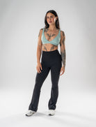 A woman modeling a two-piece activewear set consisting of a light sage green scoop-neck sports bra and black high-waisted flared leggings, standing against a light gray studio background.