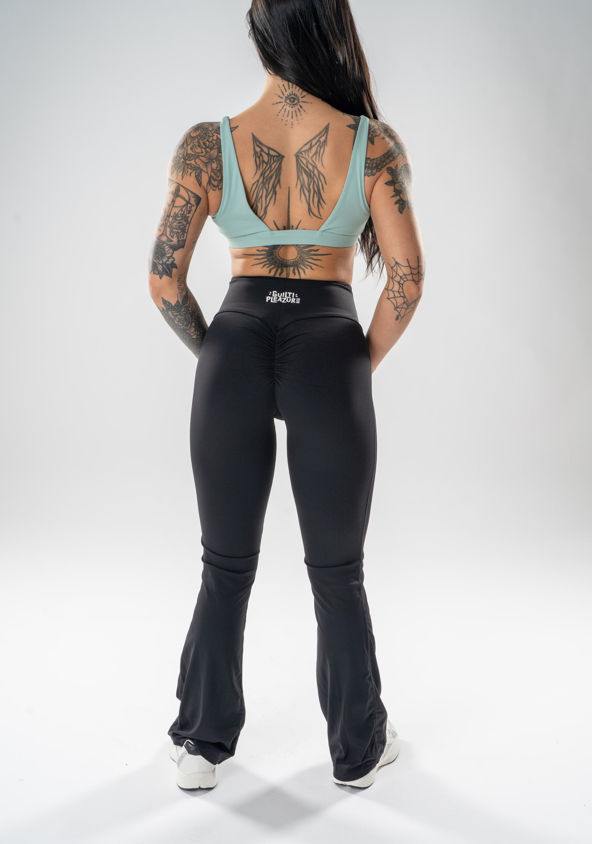 A woman viewed from the back wearing black high-waisted flared leggings and a mint green sports bra, standing against a light gray studio background.