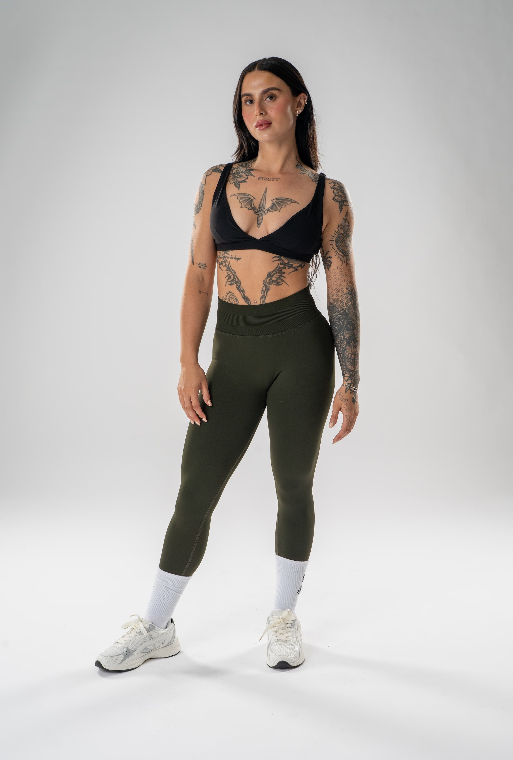 A woman modeling high-waisted olive green athletic leggings, a black sports bra, white socks, and white sneakers against a light gray studio background.