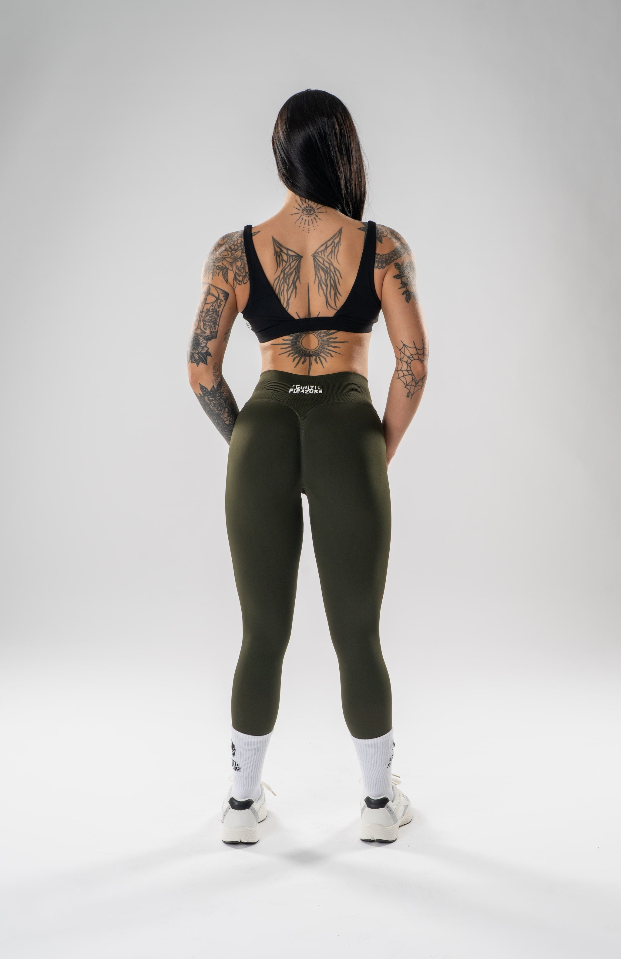 A woman stands with her back to the camera wearing olive green high-waisted leggings, a black sports bra, white socks, and white sneakers on a light gray studio background. The brand name 'altea' is visible on the waistband of the leggings.