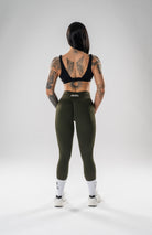 A woman stands with her back to the camera wearing olive green high-waisted leggings, a black sports bra, white socks, and white sneakers on a light gray studio background. The brand name 'altea' is visible on the waistband of the leggings.