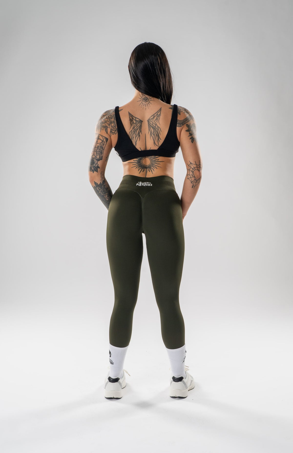 A woman stands with her back to the camera wearing olive green high-waisted leggings, a black sports bra, white socks, and white sneakers on a light gray studio background. The brand name 'altea' is visible on the waistband of the leggings.