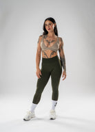 A woman modeling a matching athletic set consisting of an olive green high-waisted leggings and a taupe-beige sports bra, paired with white socks and white sneakers, against a light gray studio background.