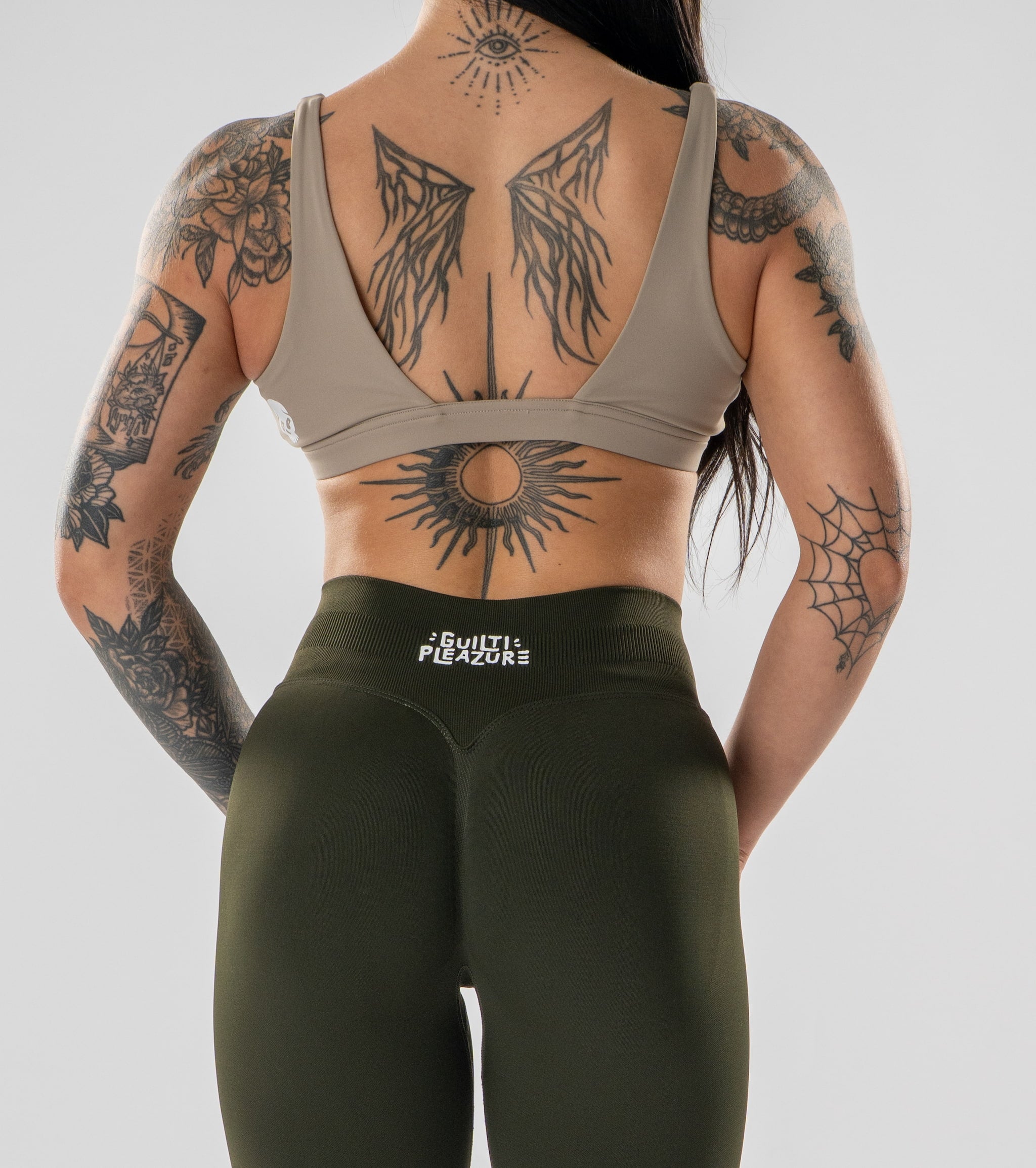 Rear view of a tattooed woman wearing a taupe sports bra and olive green leggings with the brand name 'EXCELLENT FUSION' on the waistband, against a light gray studio background.