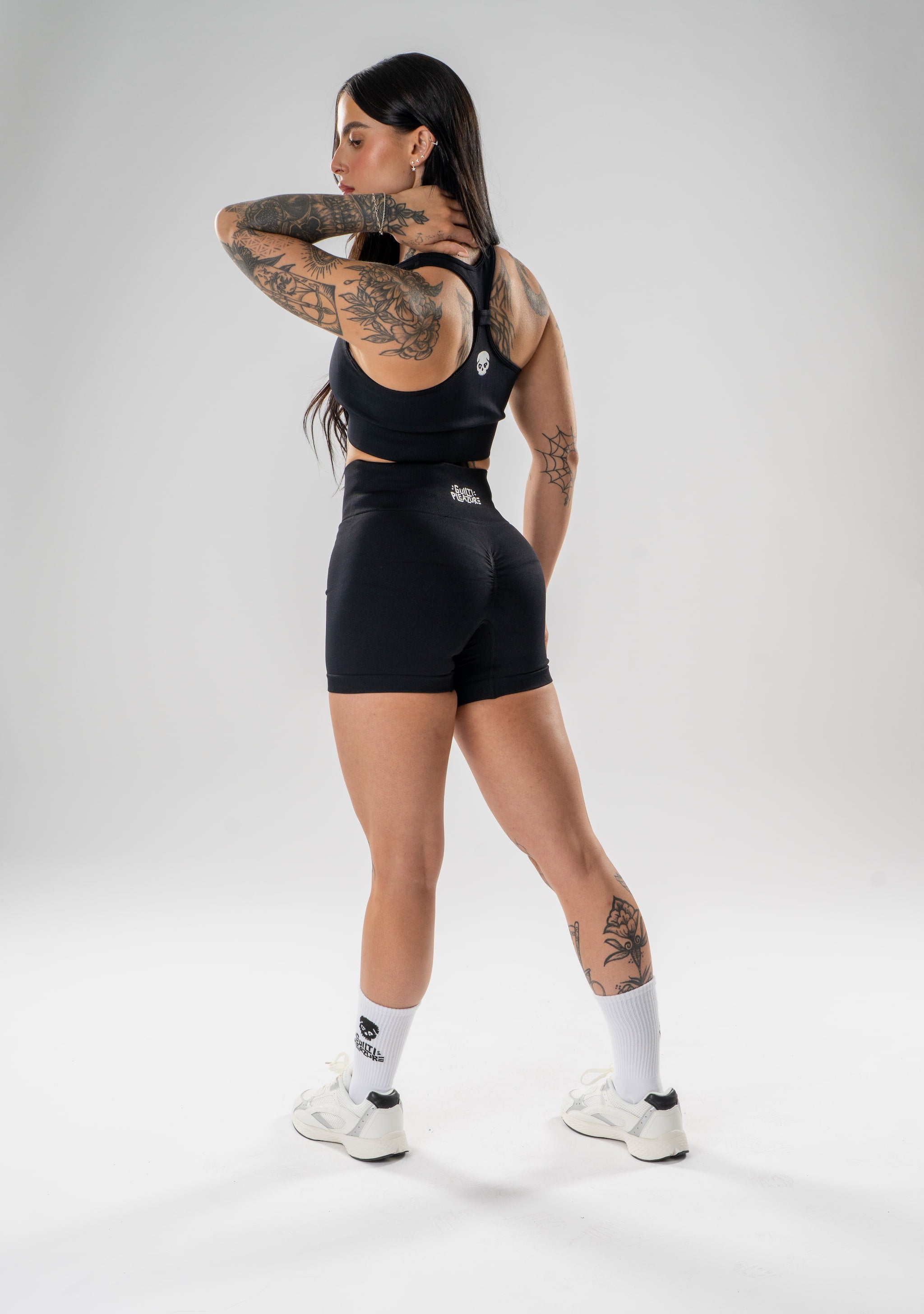 A woman modeling a black athletic set consisting of a sports bra and matching shorts, shown from a rear three-quarter angle against a light gray studio background.