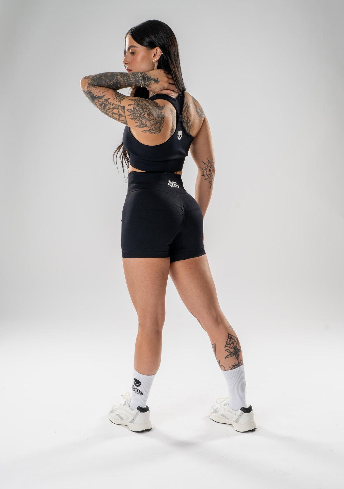 A woman modeling a black athletic set consisting of a sports bra and matching shorts, shown from a rear three-quarter angle against a light gray studio background.