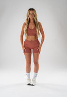 A woman modeling a matching terracotta-colored activewear set consisting of a scoop-neck sports bra and high-waisted biker shorts, paired with white crew socks and white sneakers, against a light gray studio background.