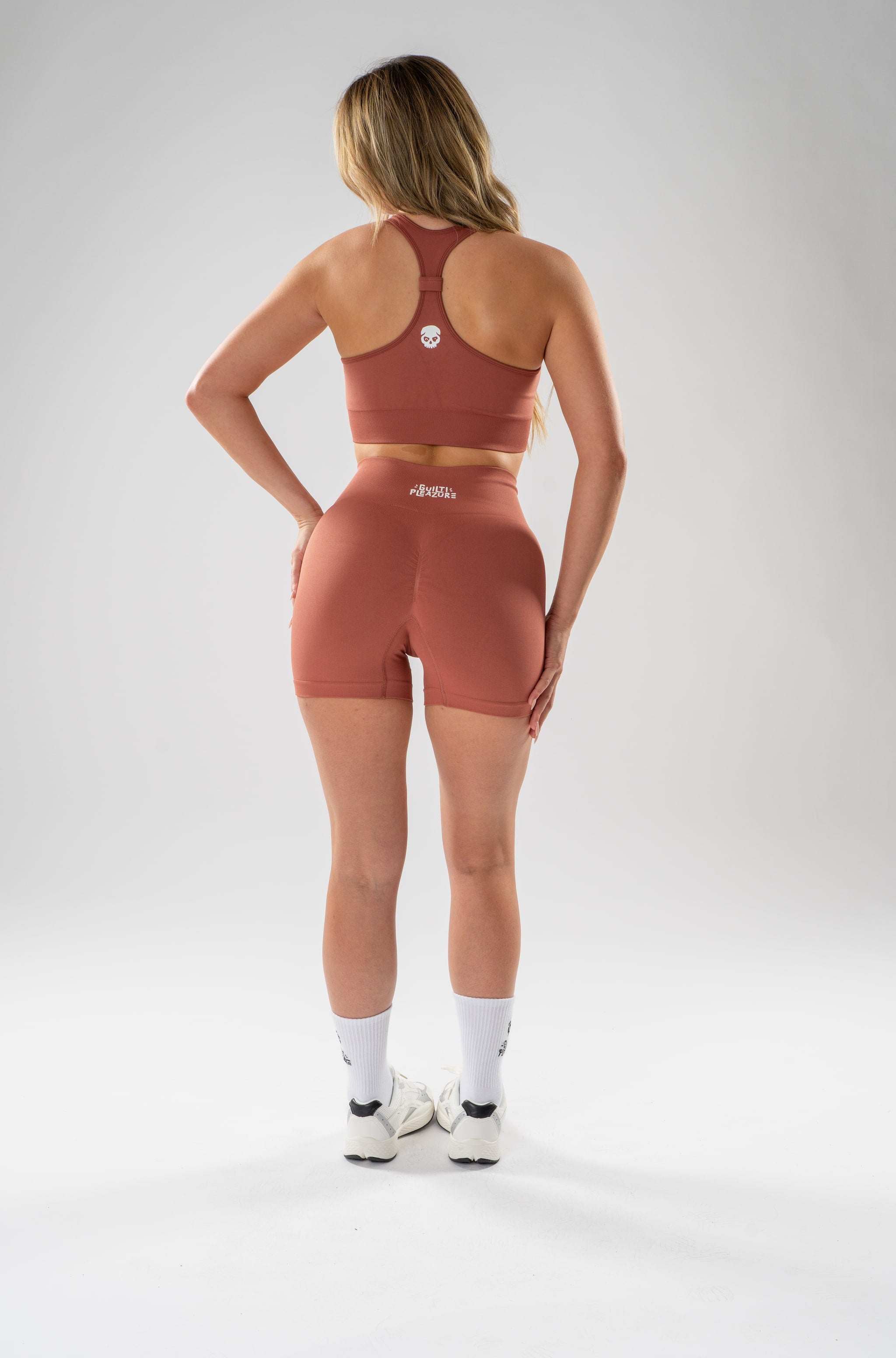 A woman stands with her back to the camera wearing a matching terracotta-colored athletic set consisting of a sports bra and high-waisted shorts, paired with white socks and sneakers, against a light gray studio background.