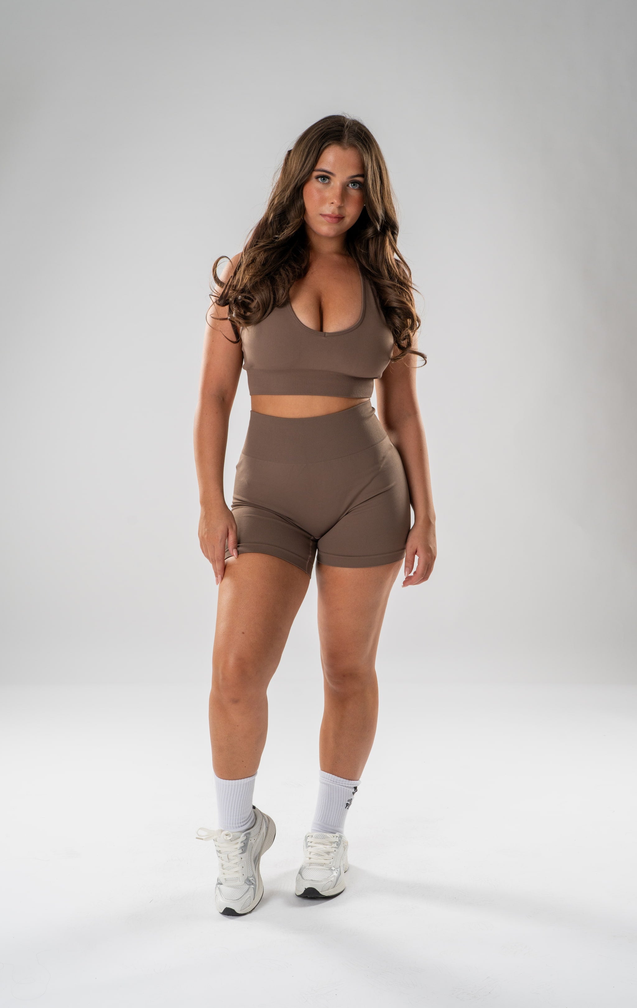 A woman modeling a matching taupe brown activewear set consisting of a scoop-neck sports bra and high-waisted shorts, paired with white socks and white sneakers, against a light gray studio background.