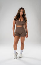 A woman modeling a matching taupe brown activewear set consisting of a scoop-neck sports bra and high-waisted shorts, paired with white socks and white sneakers, against a light gray studio background.