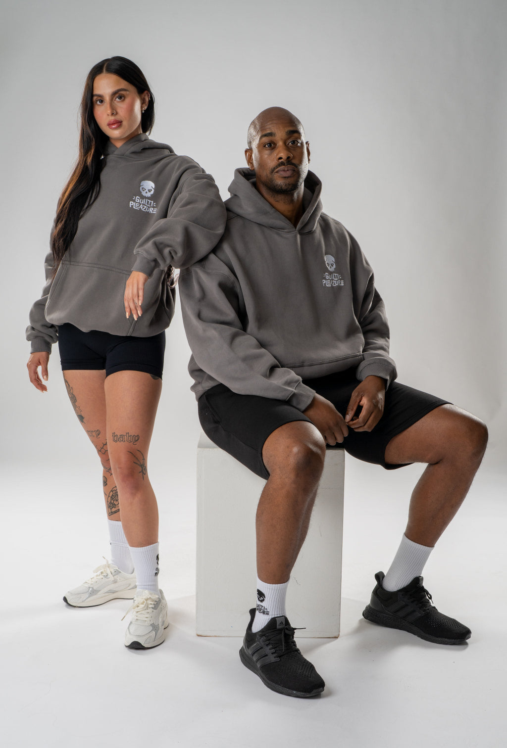 A man and a woman modeling matching dark gray hoodies with a white shield logo on the chest, paired with black athletic shorts and white or black sneakers, posed against a light gray studio background.