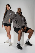 A man and a woman modeling matching dark gray hoodies with a white shield logo on the chest, paired with black athletic shorts and white or black sneakers, posed against a light gray studio background.